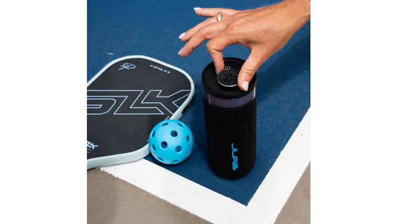 A hand adjusts the volume dial on a JLab Go Party speaker next to a pickleball paddle and ball A hand adjusts the volume dial on a JLab Go Party speaker next to a pickleball paddle and ball