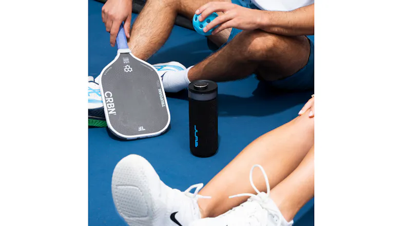 JLab Go Party speaker sitting on a blue pickleball court next to players and a paddle JLab Go Party speaker sitting on a blue pickleball court next to players and a paddle