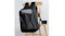A black hard‑shell backpack with a geometric front design hanging on a wooden chair, shown beside a camera A black hard‑shell backpack with a geometric front design hanging on a wooden chair, shown beside a camera