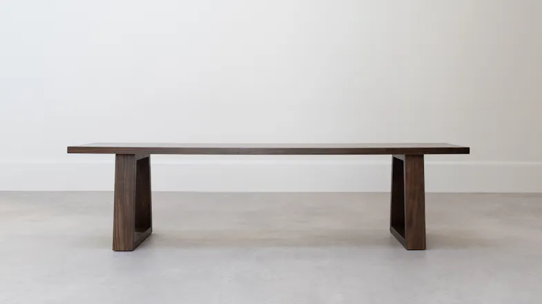 Front view of a long wooden dining bench with a rectangular seat supported by two wide, open-frame legs Front view of a long wooden dining bench with a rectangular seat supported by two wide, open-frame legs