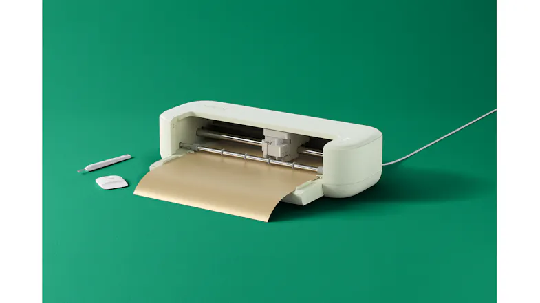Cricut Explore 5 on a green background with tools beside it and a sheet of material loaded for cutting Cricut Explore 5 on a green background with tools beside it and a sheet of material loaded for cutting