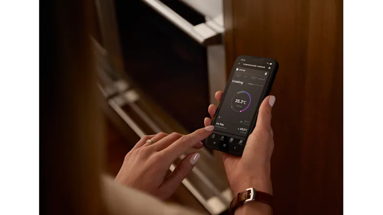 Hands holding a smartphone displaying a cooking‑monitoring app interface while near the oven Hands holding a smartphone displaying a cooking‑monitoring app interface while near the oven