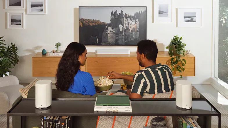 A couple on a sofa with two speakers positioned behind them A couple on a sofa with two speakers positioned behind them