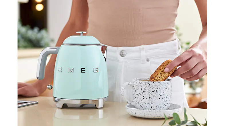 A person is dipping a biscotti into a mug next to the mini kettle A person is dipping a biscotti into a mug next to the mini kettle