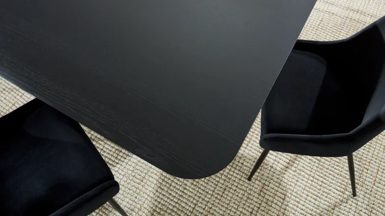 Close-up of the dark wood-look tabletop with rounded corner edges, and surrounding black dining chairs Close-up of the dark wood-look tabletop with rounded corner edges, and surrounding black dining chairs