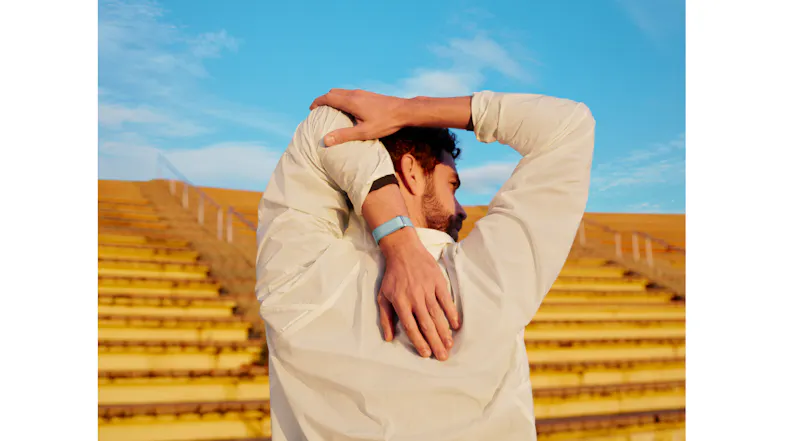 A person stretching their arms outdoors while wearing a light blue fitness band, with stadium seats and a blue sky in the background A person stretching their arms outdoors while wearing a light blue fitness band, with stadium seats and a blue sky in the background