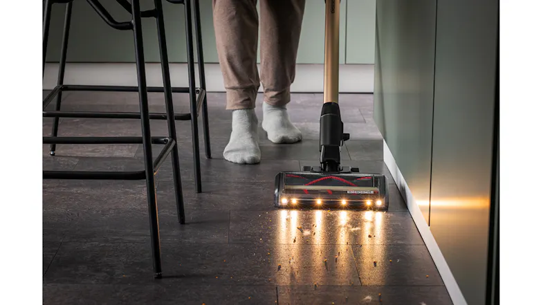 A person using the Bissell Handstick Vacuum Cleaner is cleaning crumbs along the kitchen base cabinets on a dark tile floor near stools and a sock A person using the Bissell Handstick Vacuum Cleaner is cleaning crumbs along the kitchen base cabinets on a dark tile floor near stools and a sock