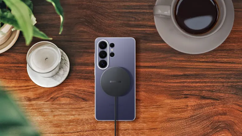 Samsung magnetic wireless charger attached to the back of a smartphone on a wooden table beside a cup and a plant, showing real‑world use Samsung magnetic wireless charger attached to the back of a smartphone on a wooden table beside a cup and a plant, showing real‑world use