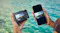 Hands holding a compact camera and a smartphone, both displaying the same marina photo over turquoise water Hands holding a compact camera and a smartphone, both displaying the same marina photo over turquoise water