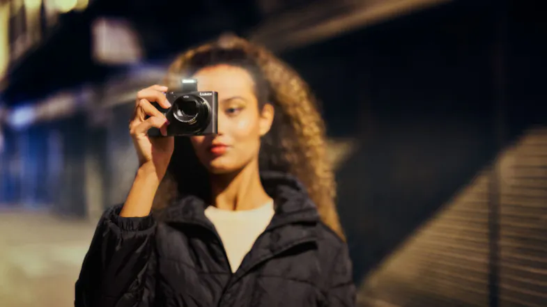 A person holding a Panasonic Lumix camera at eye level with the flash on, photographing in a dimly lit setting A person holding a Panasonic Lumix camera at eye level with the flash on, photographing in a dimly lit setting