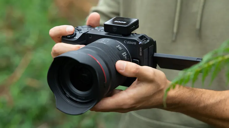 Canon mirrorless camera with lens and RODE wireless mic mounted, held outdoors with flipped screen Canon mirrorless camera with lens and RODE wireless mic mounted, held outdoors with flipped screen