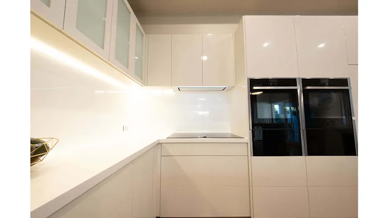 Bright kitchen with built-in ovens, white cabinetry, a stone countertop, and an integrated white glass undermount rangehood Bright kitchen with built-in ovens, white cabinetry, a stone countertop, and an integrated white glass undermount rangehood