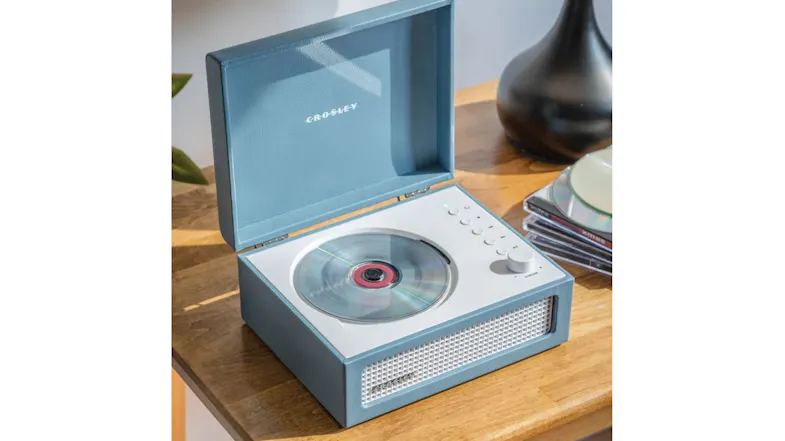 Blue Crosley CD player on a wooden table with a disc inside, control buttons visible, and a stack of CDs beside it Blue Crosley CD player on a wooden table with a disc inside, control buttons visible, and a stack of CDs beside it