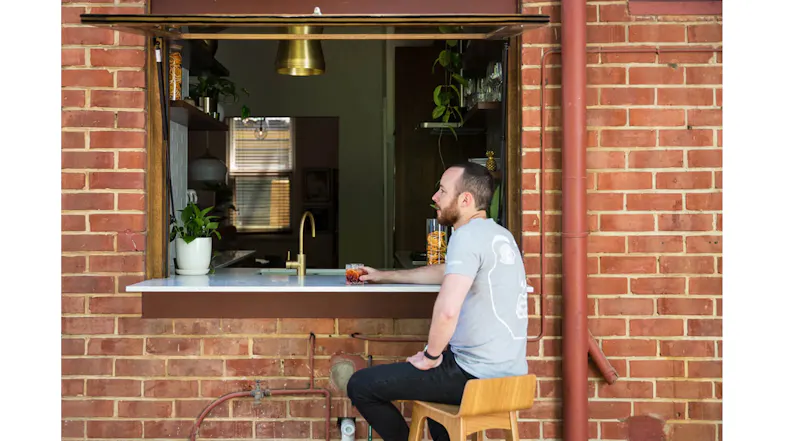 Person seated at an outdoor window bar, holding a drink, looking into a modern kitchen framed by a brick wall Person seated at an outdoor window bar, holding a drink, looking into a modern kitchen framed by a brick wall