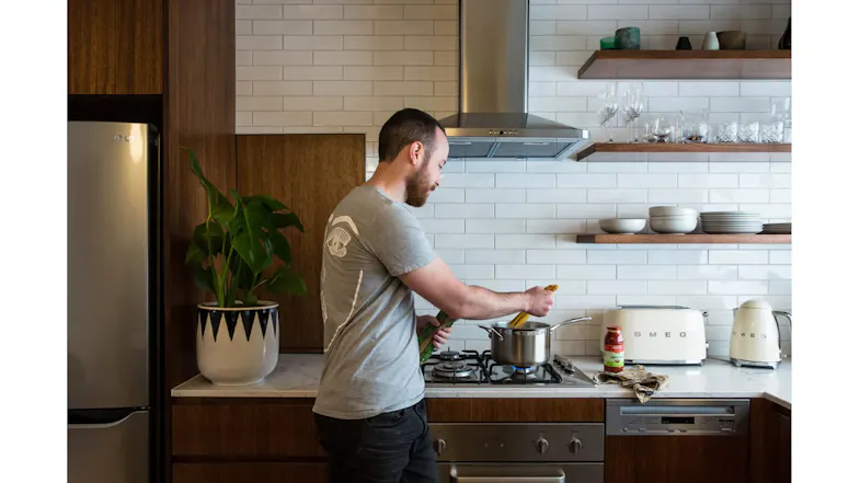 Person cooking at a stovetop beneath a stainless steel rangehood in a modern kitchen with shelves, dishes, and appliances Person cooking at a stovetop beneath a stainless steel rangehood in a modern kitchen with shelves, dishes, and appliances