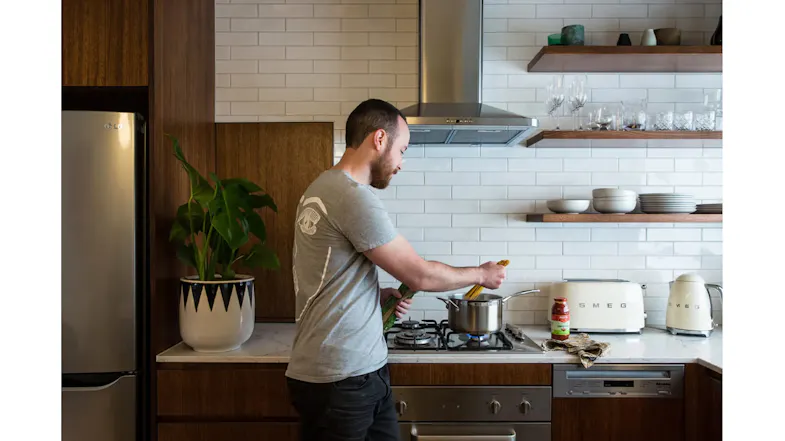 Person cooking at a stovetop beneath a stainless steel rangehood in a modern kitchen with shelves, dishes, and appliances Person cooking at a stovetop beneath a stainless steel rangehood in a modern kitchen with shelves, dishes, and appliances