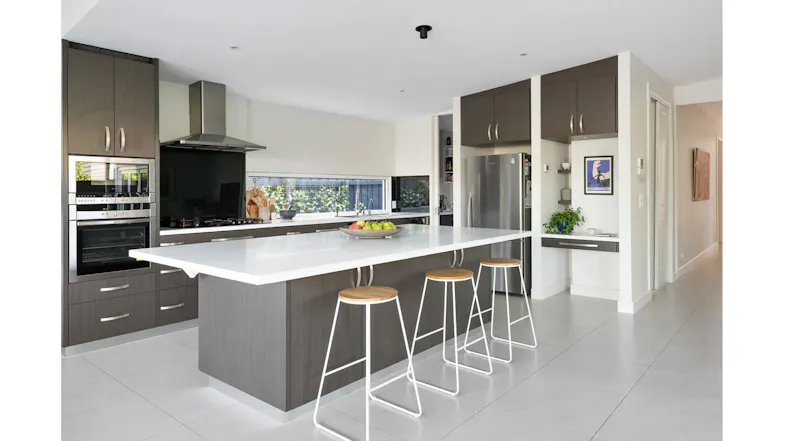 Modern kitchen with large island bench, stainless steel rangehood, dark cabinetry and integrated oven appliances Modern kitchen with large island bench, stainless steel rangehood, dark cabinetry and integrated oven appliances