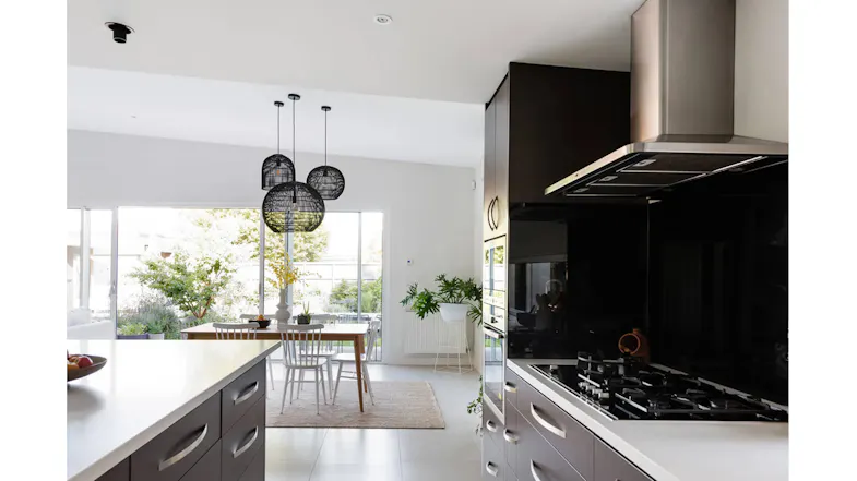 Side view of kitchen with cooktop, stainless steel rangehood, black splashback, cabinetry and dining area with garden view Side view of kitchen with cooktop, stainless steel rangehood, black splashback, cabinetry and dining area with garden view
