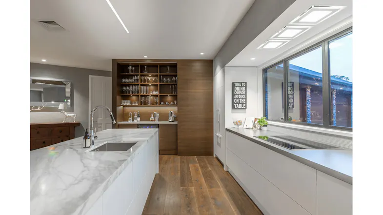 Modern kitchen with marble island, built-in sink, warm wood shelving, glassware display, large window, and sleek white cabinetry Modern kitchen with marble island, built-in sink, warm wood shelving, glassware display, large window, and sleek white cabinetry