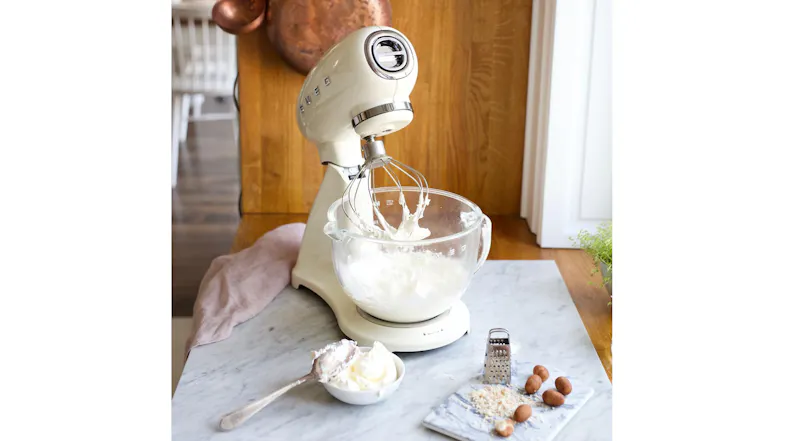 Lifestyle kitchen scene featuring the cream Smeg mixer in use with a wire whisk, mixing white cream Lifestyle kitchen scene featuring the cream Smeg mixer in use with a wire whisk, mixing white cream