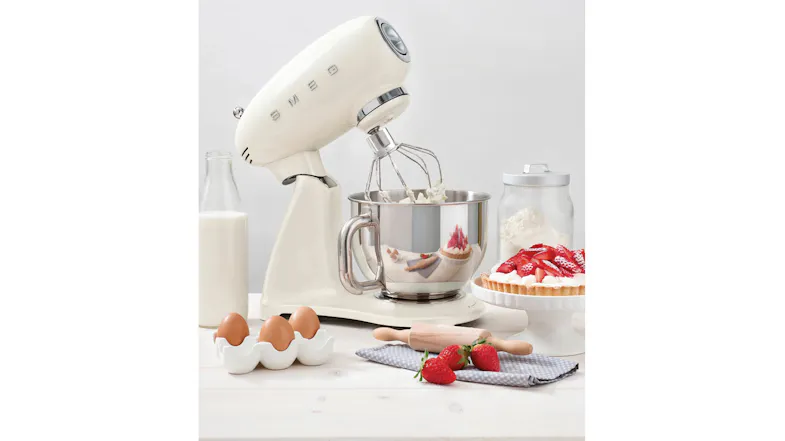 Lifestyle scene of a cream Smeg 50’s Style stand mixer with tilt head raised and wire whisk mixing cream Lifestyle scene of a cream Smeg 50’s Style stand mixer with tilt head raised and wire whisk mixing cream