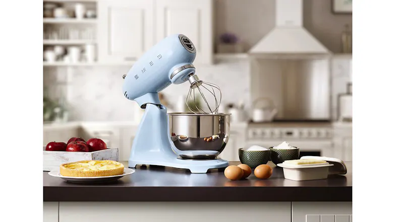 Smeg stand mixer with tilt head raised and wire whisk attachment over a stainless steel bowl on a countertop Smeg stand mixer with tilt head raised and wire whisk attachment over a stainless steel bowl on a countertop