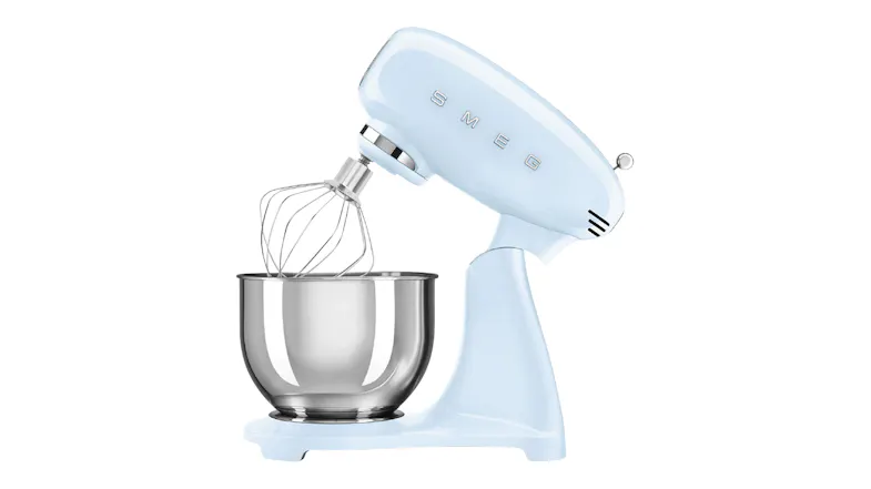 Side angle of the Smeg stand mixer with tilt head raised and wire whisk attached above a stainless steel bowl Side angle of the Smeg stand mixer with tilt head raised and wire whisk attached above a stainless steel bowl