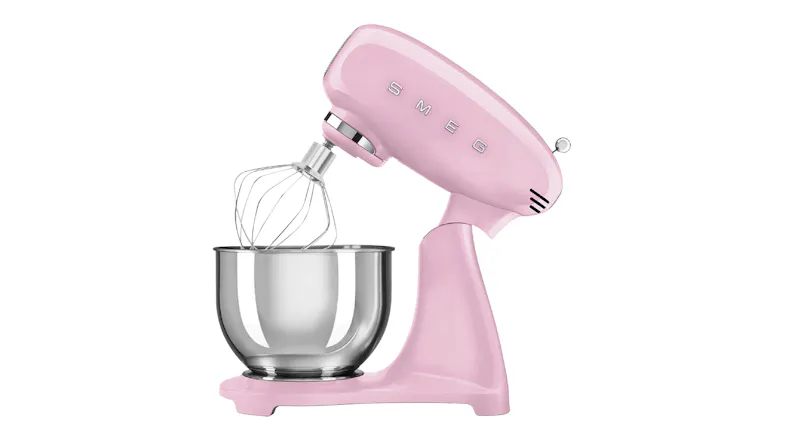 Side angle of the Smeg stand mixer with tilt head raised and wire whisk attached above a stainless steel bowl Side angle of the Smeg stand mixer with tilt head raised and wire whisk attached above a stainless steel bowl