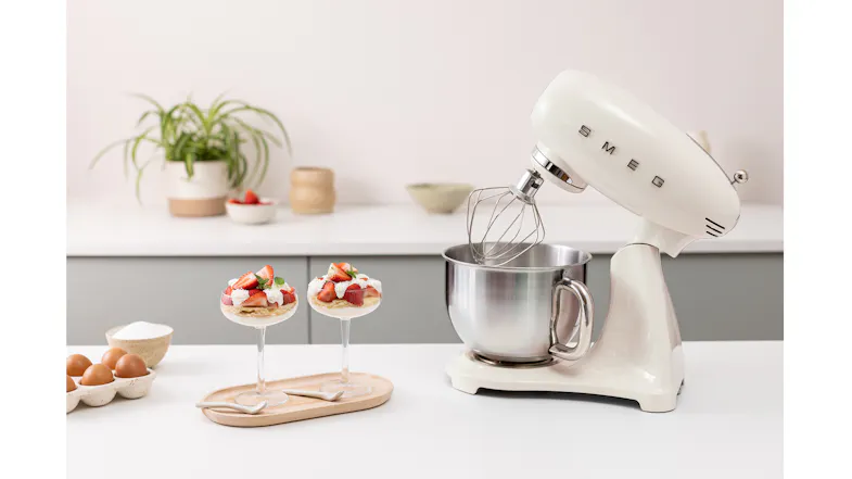 Glossy white Smeg 50’s Style stand mixer with tilt head raised and wire whisk over a stainless steel bowl Glossy white Smeg 50’s Style stand mixer with tilt head raised and wire whisk over a stainless steel bowl