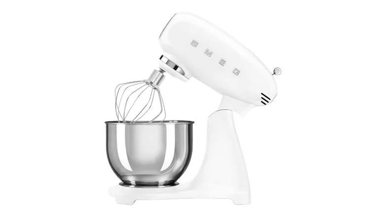 Side angle of the Smeg white stand mixer with tilt head raised and wire whisk attached above a stainless steel bowl Side angle of the Smeg white stand mixer with tilt head raised and wire whisk attached above a stainless steel bowl