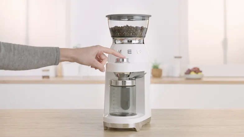 A hand pressing the start knob on a Smeg coffee grinder with beans in the hopper, placed on a wooden kitchen counter A hand pressing the start knob on a Smeg coffee grinder with beans in the hopper, placed on a wooden kitchen counter