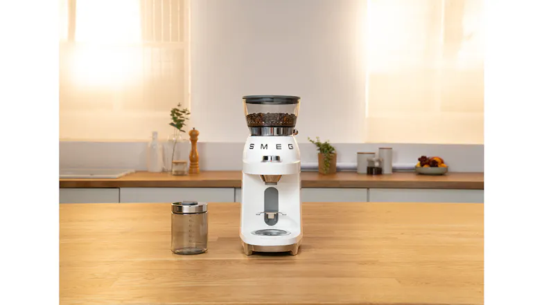 White Smeg coffee grinder on wooden kitchen counter with glass jar beside it, beans visible in hopper under warm light White Smeg coffee grinder on wooden kitchen counter with glass jar beside it, beans visible in hopper under warm light