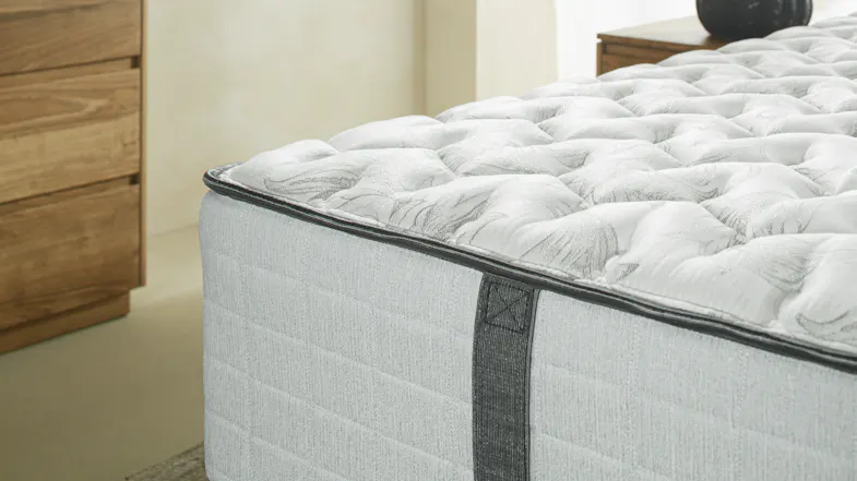 A close-up of a mattress corner showing a tufted white top, grey side panels, and a stitched handle beside a wooden dresser A close-up of a mattress corner showing a tufted white top, grey side panels, and a stitched handle beside a wooden dresser
