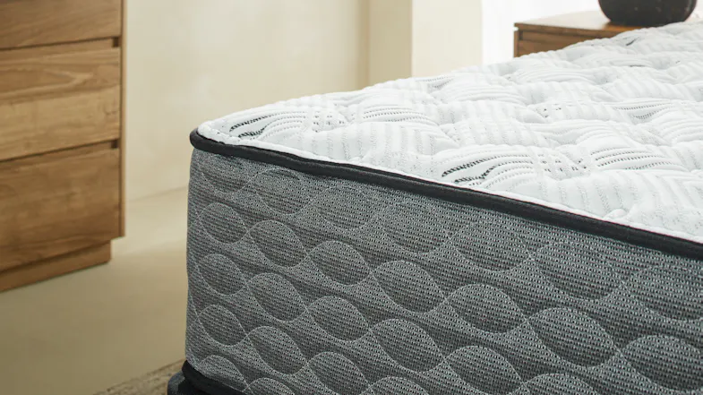 Close-up of a Serene mattress corner with a white quilted top surface and a dark grey, wave-patterned textured side fabric Close-up of a Serene mattress corner with a white quilted top surface and a dark grey, wave-patterned textured side fabric