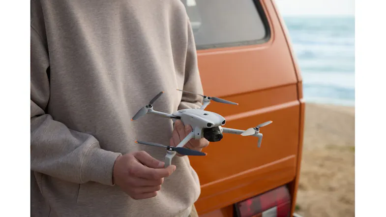 A hand holding a compact foldable camera drone beside an orange vehicle near the beach A hand holding a compact foldable camera drone beside an orange vehicle near the beach