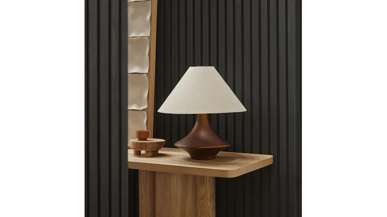 Angled view of the lamp on a wooden table, showcasing a bronze base shape and a softly flared fabric shade Angled view of the lamp on a wooden table, showcasing a bronze base shape and a softly flared fabric shade
