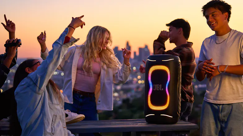 The speaker placed on a table while a bunch of people gathers around to listen to music The speaker placed on a table while a bunch of people gathers around to listen to music