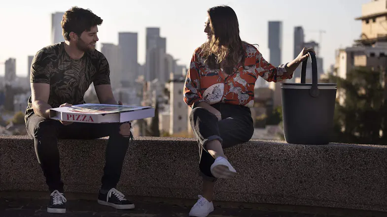 Two people are seated on a rooftop ledge with a pizza box and a black portable Keter cooler against a city skyline Two people are seated on a rooftop ledge with a pizza box and a black portable Keter cooler against a city skyline