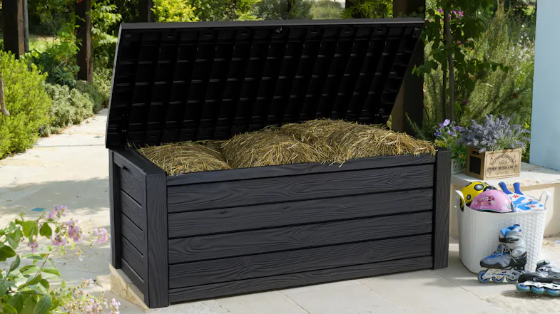 Black Keter Eastwood outdoor storage box with an open lid, filled with straw, on a garden patio surrounded by plants Black Keter Eastwood outdoor storage box with an open lid, filled with straw, on a garden patio surrounded by plants