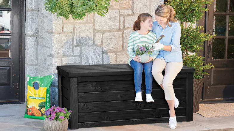 Two people sit on a black outdoor storage bench beside potted flowers and gardening supplies Two people sit on a black outdoor storage bench beside potted flowers and gardening supplies