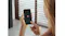 A woman holding a smartphone displaying the red Miele app icon on a black screen, with blurred window blinds in the background A woman holding a smartphone displaying the red Miele app icon on a black screen, with blurred window blinds in the background