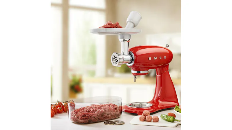Multi food grinder fitted to a red Smeg stand mixer in a bright kitchen setting. Meat and ingredients on the countertop Multi food grinder fitted to a red Smeg stand mixer in a bright kitchen setting. Meat and ingredients on the countertop