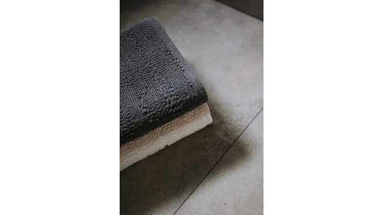 Close-up of folded textured Baksana Lisbon bath mats in Slate, Linen, and White on a tiled bathroom floor Close-up of folded textured Baksana Lisbon bath mats in Slate, Linen, and White on a tiled bathroom floor