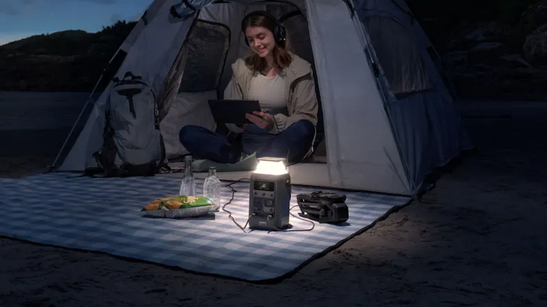 A camper using a tablet in a tent with a portable power station and an LED lantern for off-grid power A camper using a tablet in a tent with a portable power station and an LED lantern for off-grid power