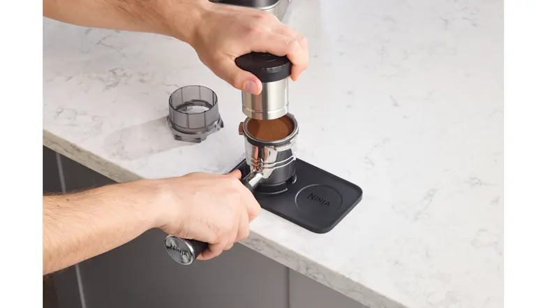 Person holding a tamper over a portafilter resting on a black Ninja tamping mat Person holding a tamper over a portafilter resting on a black Ninja tamping mat