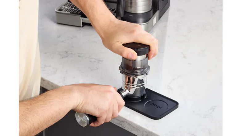 Person pressing down on a tamper with a funnel attached to a portafilter on a Ninja mat Person pressing down on a tamper with a funnel attached to a portafilter on a Ninja mat