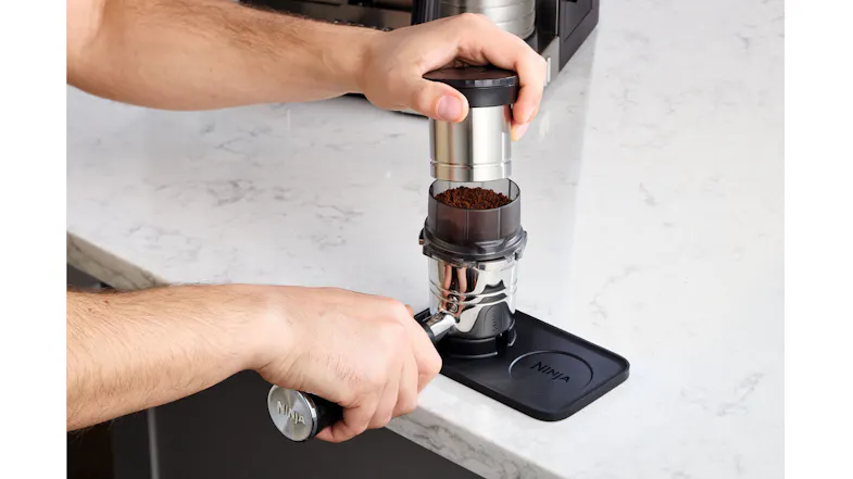 Hands aligning a metal tamper over a funnel-equipped portafilter on a silicone mat Hands aligning a metal tamper over a funnel-equipped portafilter on a silicone mat