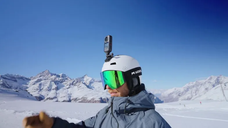 Snowboarder wearing a white helmet with a 360-action camera mounted on top against snowy mountains Snowboarder wearing a white helmet with a 360-action camera mounted on top against snowy mountains