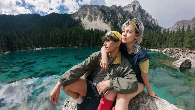Couple sitting together on a rock beside a vibrant turquoise mountain lake surrounded by pine trees Couple sitting together on a rock beside a vibrant turquoise mountain lake surrounded by pine trees