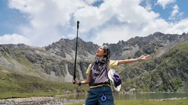 Hiker standing in a grassy mountain valley holding an extended selfie stick with a 360-action camera Hiker standing in a grassy mountain valley holding an extended selfie stick with a 360-action camera